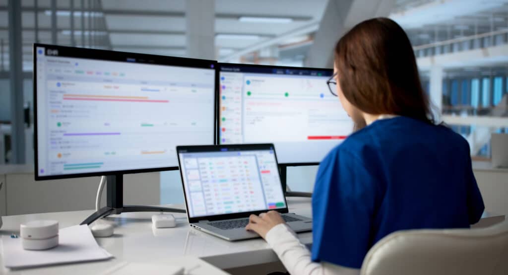 Healthcare IT professional working on a laptop with dual monitors to review digital records and asset tracking.